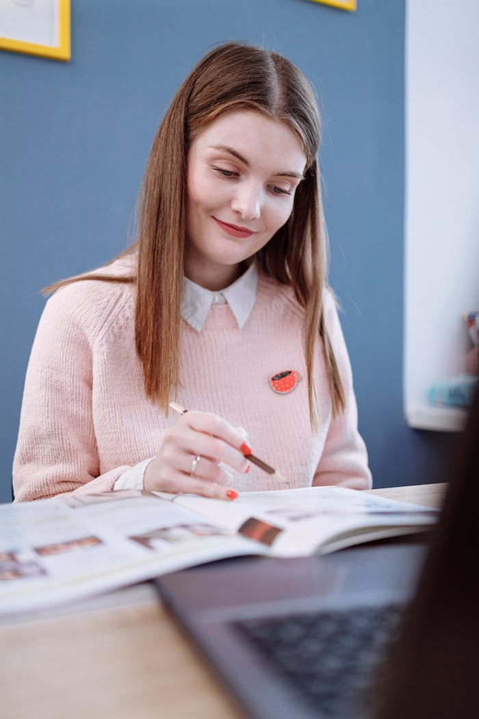 A young woman in a cozy pink sweater focuses on studying at a table with books and a laptop.